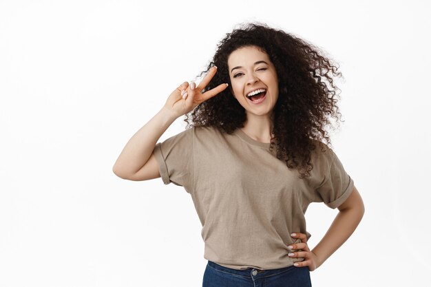 woman, laughing and smiling, showing peace v-sign near eye, kawaii pose, standing on white