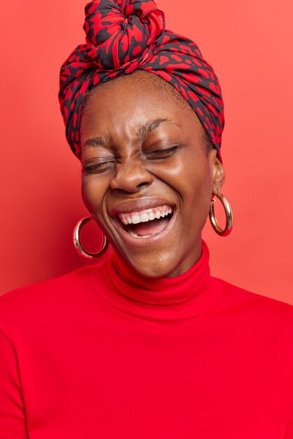 woman laughs gladfully has carefree expression wears scarf on head turtleneck and earrings isolated on red. Happy emotions concept