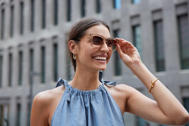 женщина искренне смеется, одетая в модную одежду, модные солнцезащитные очки с широкой улыбкой на лице проводит свободное время, гуляя по городу