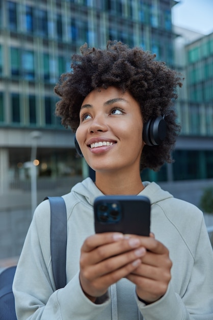 woman leads sporty lifestyle listens favorite music in wireless headphones holds smartphone downloads songs to playlist focused somewhere prepares for workout training