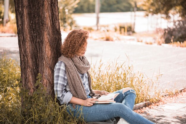 Woman leaning on tree and reading book in public garden