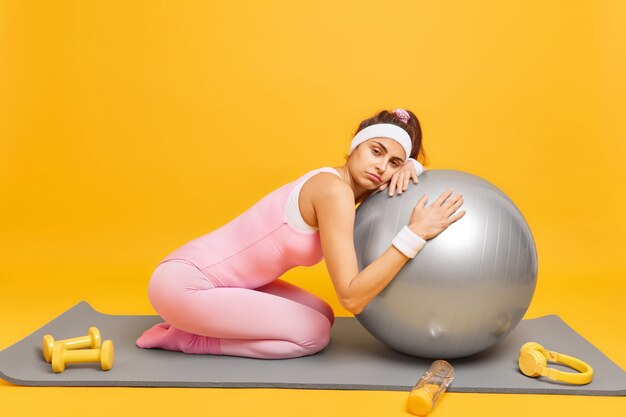 woman leans at fitness ball feels fatigue after aerobic training wears headband wristband and activewear poses on karemat isolated on yellow 