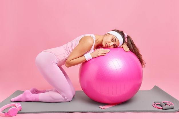 woman leans at fitness ball has tired expression trains at home gym dressed in activewear poses on knees 