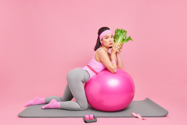 woman leans at swiss ball holds green vegetable eats healthy food leads active lifestyle has regular workout to be fit poses on fitness mat