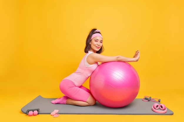 woman lenas at fitness ball has satisfied expression dressed in activewear takes break after training at home likes gymnastics and aerobics poses on mat indoor