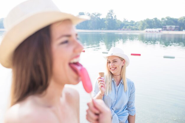 湖の近くにアイスクリームコーンを持っている彼女の友人の前でポピーを舐める女性