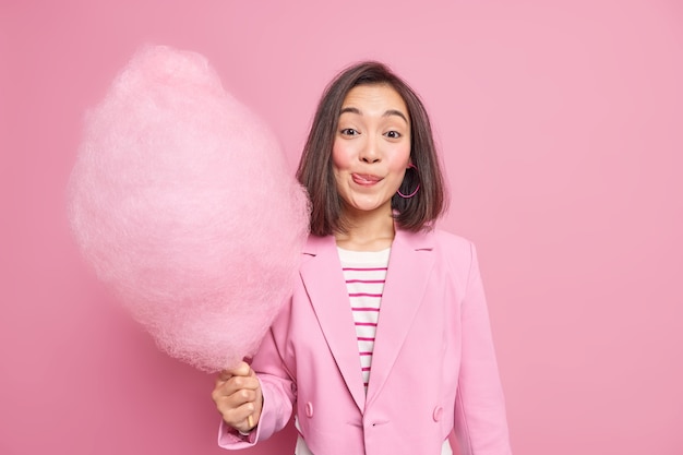 woman licks lips as holds appetizing sweet candy floss dressed in fashionable formal outfit 