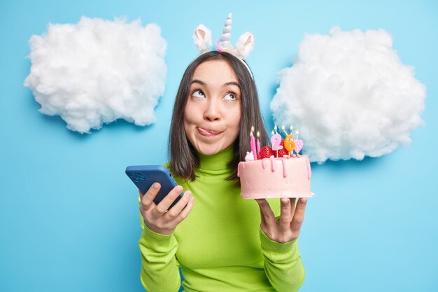 woman licks lips holds delicious cake with burning candles celebrates 26th birthday receieves message of congratulations on mobile phone stands