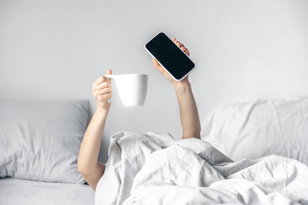 Free photo a woman lies in bed with a smartphone and a cup of coffee in her hands