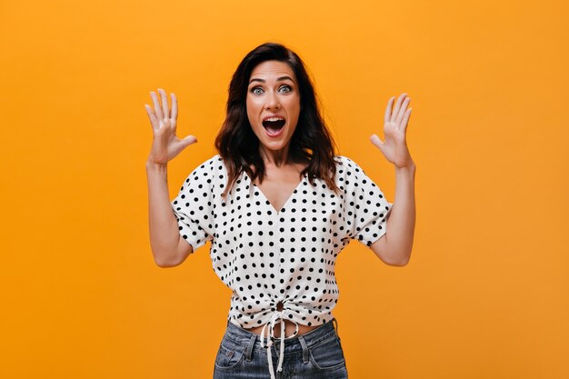 Woman in light blouse screams happily and looks into camera on orange background. Wonderful adult girl in polka dot shirt and jeans is very surprised.