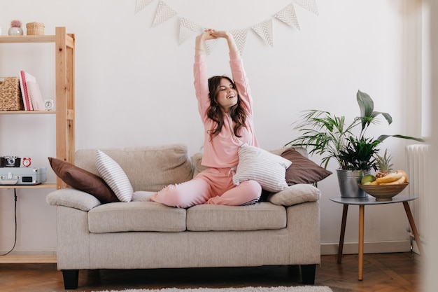 женщина в светло-розовой пижаме поднимает руки вверх после хорошего сна и позирует в квартире