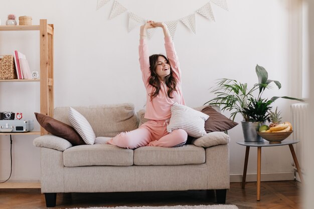 woman in light pink pajamas raises her hands up after good sleep and poses in apartment