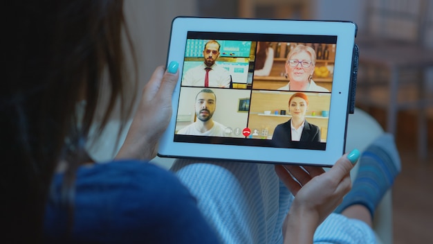 Woman listening online training on tablet late at night sitting on sofa. Remote worker having virtual meeting consulting with colleagues on videocall and webcam chat using internet technology.