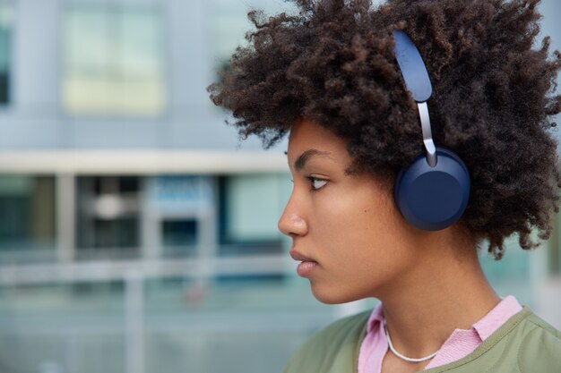 woman listens music in headphones enjoys interesting plot of audio book looks seriously forward walks in modern urban setting