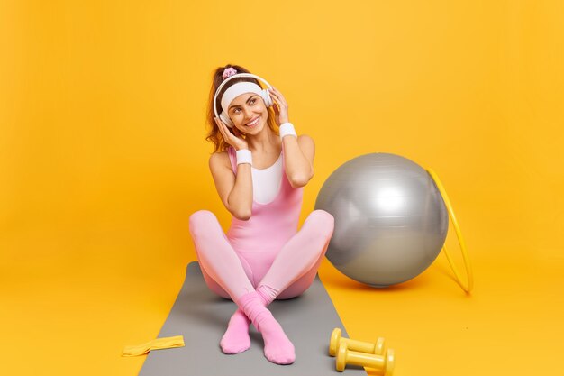 женщина слушает музыку в наушниках сидит, скрестив ноги на фитнес-коврике, делает гимнастические тренировки с обручем со швейцарским мячом и гантелями, изолированными на желтом