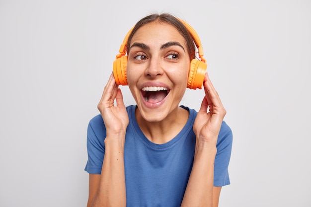 woman listens music via orange headphones dressed in casual t shirt giggles optimistic enjoys good sound on white