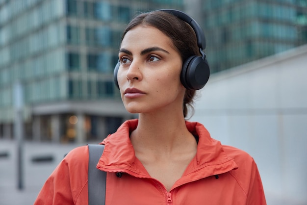 woman listens music in wireless headphones enjoys audio track dressed in anorak waits for coach poses outdoors