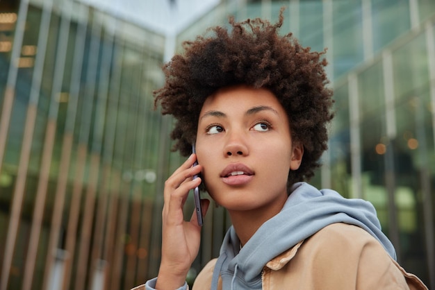 женщина слушает полученную информацию со смартфона во время разговора по телефону, одетая в толстовку с капюшоном, задумчиво смотрит в сторону, стоит на фоне затуманенного стеклянного здания города. Коммуникация