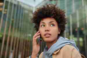 Free photo woman listens received smartphone information during cell conversation dressed in hoodie looks thoughtfully away stands against blurred glass city building. communication