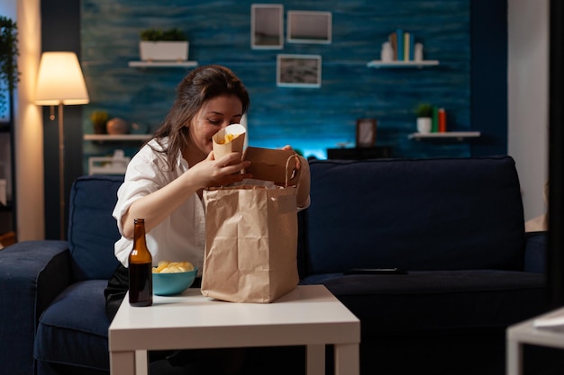 Woman in living room preparing for couch dinner unpacking french fries and tasty hamburger from takeaway paper bag. Person having burger junk food meal and bottled beer sitting on sofa.