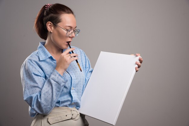 Woman looking at empty canvas and brush on gray background