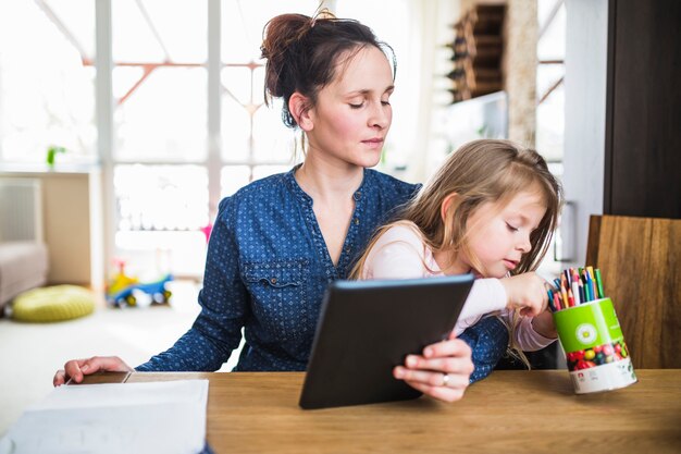 Woman looking at her daughter choosing pencil while holding digital tablet