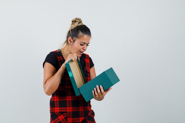 Woman looking into present box in pinafore dress and looking curious , front view.