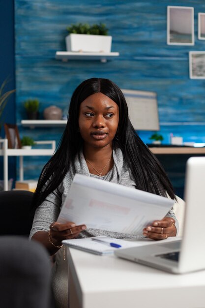 Женщина смотрит на экран ноутбука и держит документы с диаграммами данных, делает удаленную работу. Молодой человек работает дома над бизнес-проектом, используя компьютер и создавая файлы.