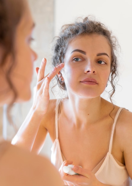 Женщина смотрит в зеркало и наносит крем на лицо