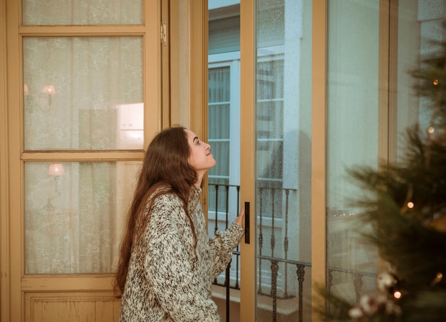 Woman looking out of window next to Christmas tree