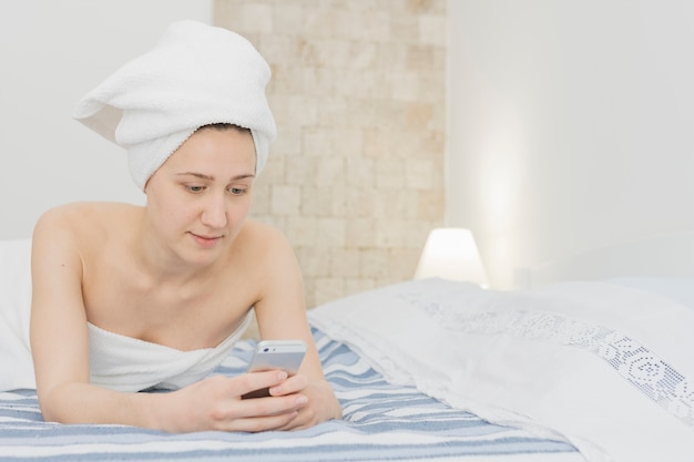 Foto gratuita donna che esamina smartphone nel letto dopo la doccia