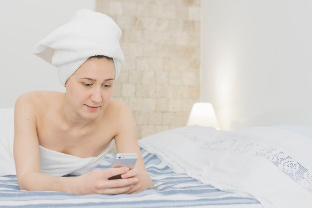 Женщина смотрит на смартфон в постели после душа