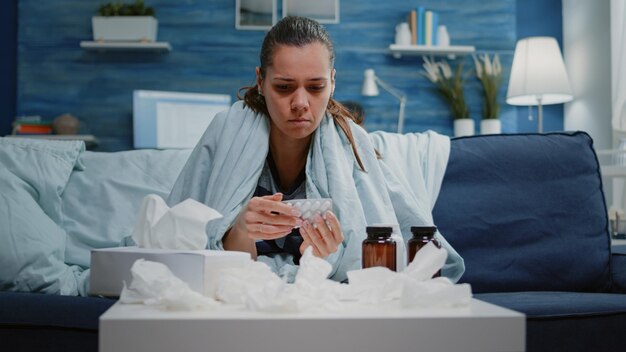 Woman looking at tablets of capsules and jar of pills on table finding healing treatment against headache and seasonal cold. Unhappy person with sickness reading medicine labels