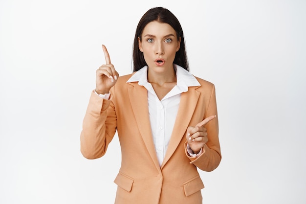 Woman looks amazed while pointing at two objects showing chart or advertisements standing in suit against white background