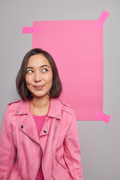 La donna distoglie lo sguardo vestita in giacca posa su grigio con foglio di carta rosa intonacato