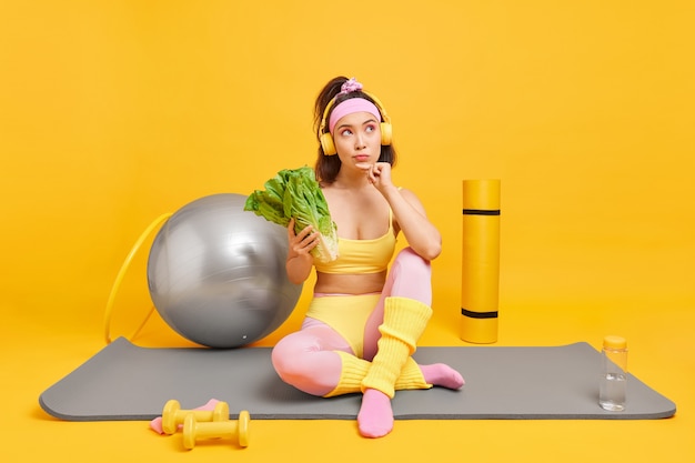 женщина смотрит в сторону, думает, глубоко держит зеленые овощи, придерживается диеты, принимает позы для здорового питания на коврике для фитнеса, использует различное спортивное оборудование для тренировок