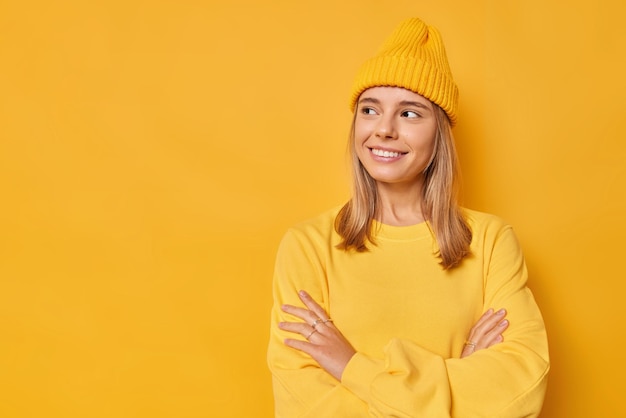 женщина смотрит в сторону с радостным выражением лица, скрестив руки, задумчиво смотрит, носит повседневный джемпер и шляпу, выделенную на ярко-желтом фоне с копией пространства