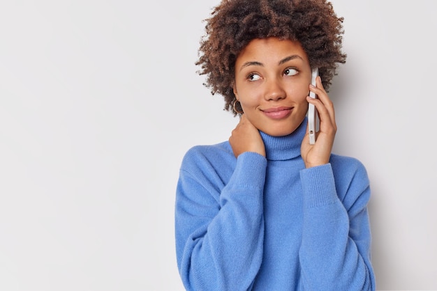 woman looks away with satisfied expression makes telephone call dressed in casual blue turtleneck isolated on white copy space for advert