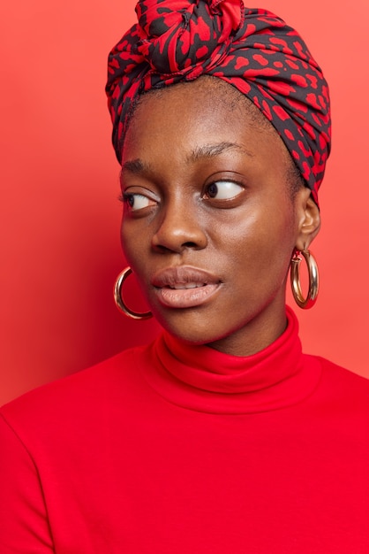  woman looks away with thoughtful expression wears turtleneck scarf tied on head big earrings poses on bright red 