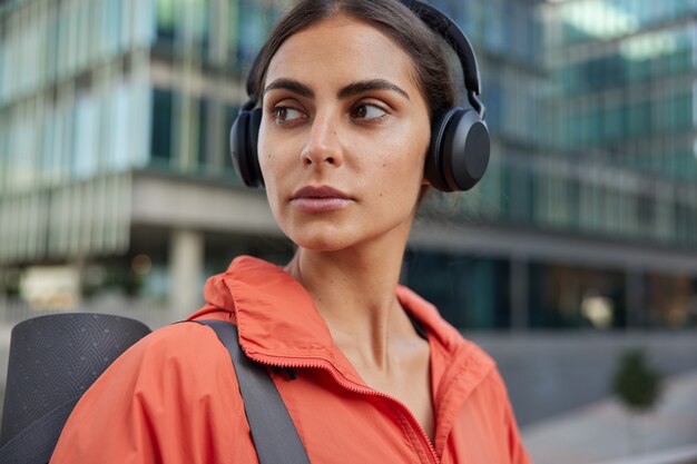  woman looks back returns from fitness training listens music via headphones wears red anorak carries rolled karemat poses against blurred city 