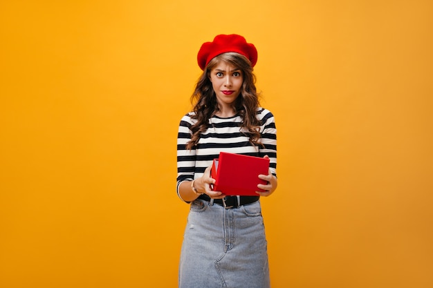 Woman looks dissatisfied after opening red box. Sad girl with curly hair in red beret and denim skirt with belt posing on isolated background.