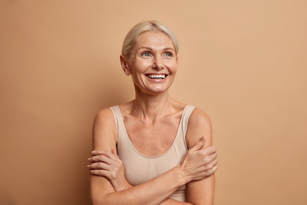 La donna guarda con gioia sopra tiene le braccia foded ha una carnagione ben curata pelle sana denti bianchi isolati su marrone