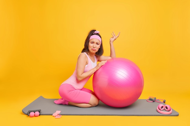 Женщина возмущенно смотрит в камеру, опирается на фитнес-мяч, регулярно тренируется дома, носит спортивную одежду, выделенную на желтом