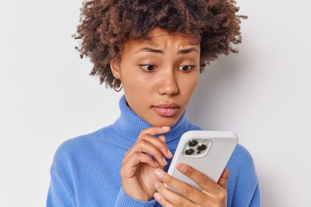 woman looks at smartphone screen with concerned expression reads bad news wears casual blue turtleneck isolated on white has complicated life