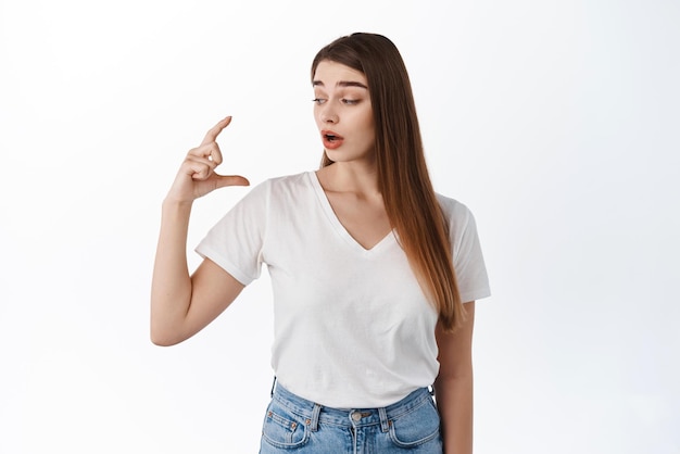 Woman looks surprised at her hand showing small little size thing raise eyebrows in surprise and disbelief holding tiny object over copy space white background