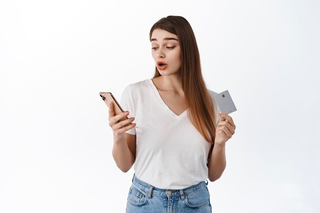 Женщина выглядит удивленной на свой смартфон, расплачиваясь кредитной картой, делая покупки в Интернете, задыхаясь и поднимая брови, заинтригованная чем-то на экране мобильного телефона, стоя над белой стеной