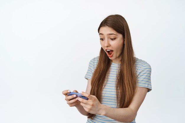 La donna sembra sorpresa dallo schermo del telefono cellulare. la ragazza reagisce a smth impressionante sullo smartphone, guardando video sul cellulare su bianco.