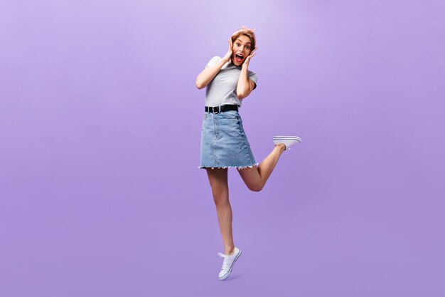 Woman looks surprised on purple background. Wonderful joyful girl in trendy outfit and white sneakers posing.n isolated backdrop.