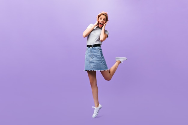 Женщина выглядит удивленной на фиолетовом фоне. Замечательная радостная девушка в модном наряде и белых кроссовках позирует на изолированном фоне.
