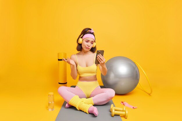 woman looks with clueless expression at smartphone display takes break after aerobics or fitness training leads healthy lifestyle dressed in sportswear poses on mat in full length